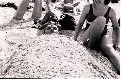 Black-and-white beach scene from the 1960s–70s, featuring relaxed beachgoers in swimwear. A man lies buried in sand up to his...