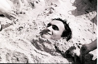 Black-and-white candid of a man buried in sand, partially buried up to his neck, with only his head and shoulders visible. Th...