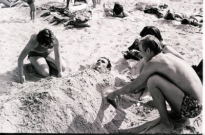 Black-and-white beach scene from the 1960s–70s, featuring relaxed beachgoers in swimwear. Three individuals kneel in sand, wh...