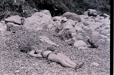 Mid-20th century beach scene: woman in leopard-print swimsuit lies on pebbled shore, arms stretched. Another figure in hat si...
