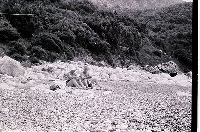 Two individuals in vintage swimwear sit on a rocky, pebble-covered shore, mid-20th century. Dense forest and rugged terrain f...