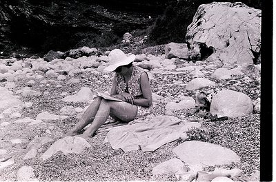 Mid-century outdoor scene: Woman in wide-brimmed hat and sleeveless dress reads on a blanket amid rocky terrain, likely a coa...