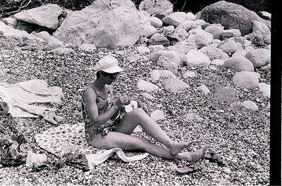 Mid-century beach scene featuring a woman in a floral swimsuit and straw hat, seated on a towel among smooth river rocks. Her...
