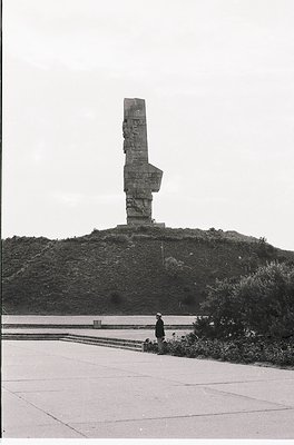 Monumental concrete slab with a raised "V" symbol atop a grassy hill, likely a Soviet-era victory memorial. Two individuals i...