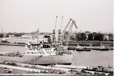 Mid-20th century industrial port scene featuring a large cargo ship docked alongside cranes and smaller vessels. Distinctive ...