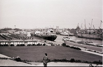 Black-and-white port scene featuring mid-20th century maritime infrastructure. Docked cargo ships, cranes, and industrial bui...