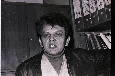 Black-and-white portrait of a man in a turtleneck sweater and leather jacket, standing indoors near stacked audio cassettes a...