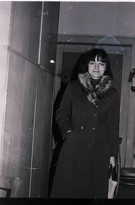 Black-and-white portrait of a woman in a long fur-collared coat, standing indoors near a frosted glass door. The coat’s butto...