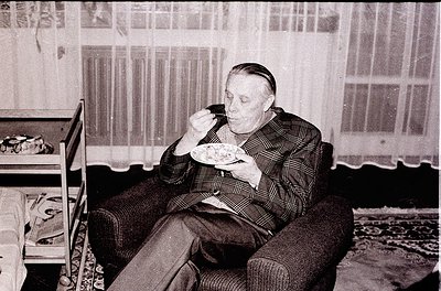 Mid-century man in plaid suit eats from a bowl while seated in a vintage armchair, suggesting 1950s–1960s domestic life. Curt...