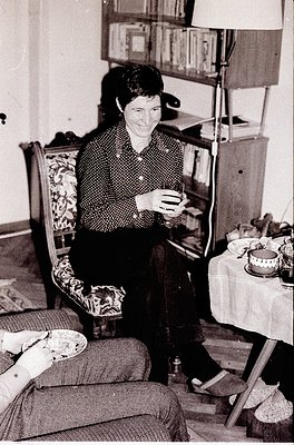 Vintage black-and-white interior shot of a woman seated on a patterned sofa, holding a small object (likely a cigarette or pi...