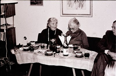Three elderly women seated around a table set with vintage glassware, plates, and a bottle of wine, likely mid-20th century. ...