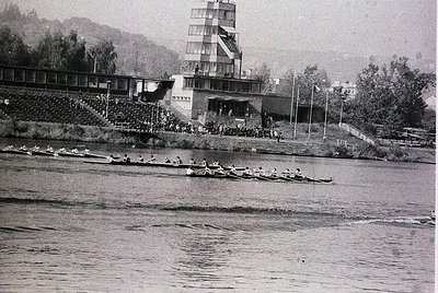 Black-and-white shot of a rowing competition at a stadium-style lakeside venue, featuring tiered concrete seating and a multi...