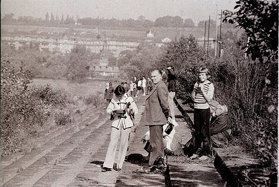 Group of people in mid-20th-century attire clearing a rural pathway, likely post-WWII. Distinctive 1950s-60s clothing: men in...