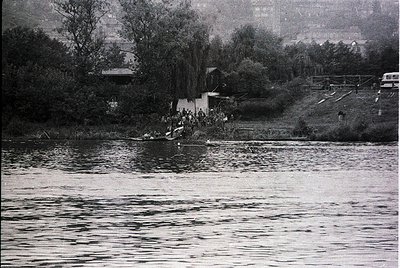 Black-and-white shot of a crowded riverside gathering, likely mid-20th century. A dense group of people stands on a rocky sho...