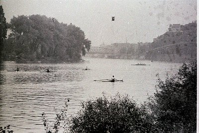 Vintage sepia-toned rower on calm river with bridge and trees in background, likely early 20th century. Wooden sculling boat ...