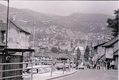 Black-and-white street scene in a mountainous European town, likely –70s. Narrow road flanked by parked vintage cars, pedestr...