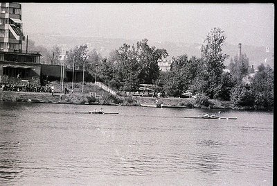 Black-and-white shot of a riverbank scene with rowers practicing in the mid-20th century. Dense greenery frames a rowing lane...