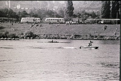 Vintage black-and-white scene of rowers competing on a calm river, with vintage buses and spectators lining the shore. Mid-20...