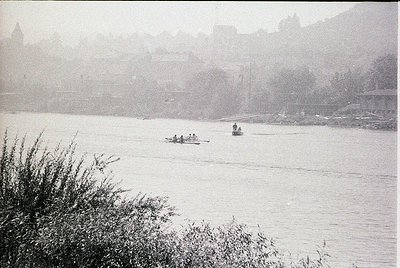 Vintage black-and-white river scene with misty urban backdrop. Small rowboats and a motorboat navigate calm waters, framed by...