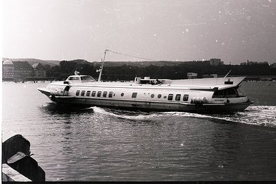 Mid-20th century ferry with sleek, elongated hull and circular portholes cruising on calm waters. Urban shoreline with brick ...