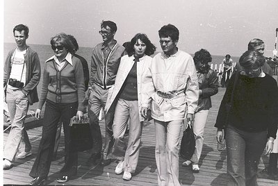 Group of seven individuals in 1970s beachwear strolling on a wooden pier, likely a seaside resort. Men wear wide-legged trous...