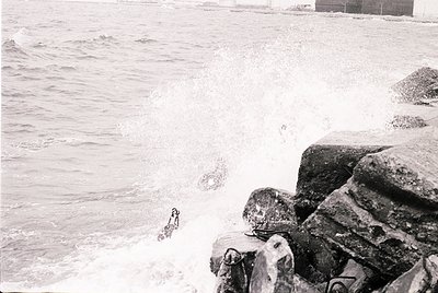 Black-and-white coastal scene featuring rough waves crashing against jagged rocks. A lone figure in mid-dive enters the water...