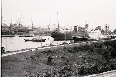 Black-and-white port scene featuring mid-20th century industrial maritime activity. Large cargo ships docked alongside cranes...