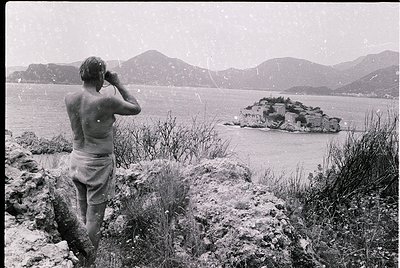 A man in mid-20th-century swimwear poses on rocky terrain, facing a rugged island fortress in a coastal bay. Snow-capped moun...