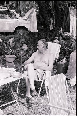 Mid-20th century man lounges in a vintage deck chair beside a vintage car and camping setup. Rock formations and a makeshift ...