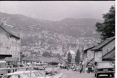 Vintage black-and-white street scene in a mountainous European town, likely 1950s–1970s. Wooden buildings with sloped roofs l...
