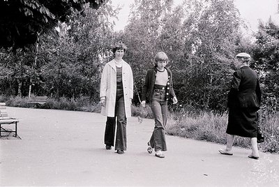Three individuals stroll along a tree-lined park path in 1970s fashion: flared jeans, striped tops, and layered cardigans. Lu...
