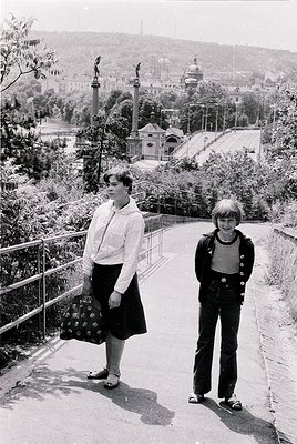 Two individuals pose on a sunlit urban pathway, likely mid-20th century (1960s–70s). The woman wears a knee-length floral-pat...