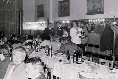 Formal banquet hall with long tables draped in white linen, featuring wine bottles, glasses, and place settings. Attendees in...