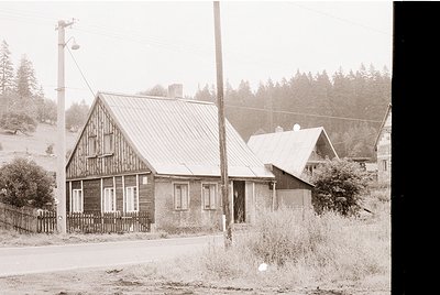Rustic wooden houses with gabled roofs in a rural setting, likely early-to-mid 20th century. Wooden picket fence and overgrow...