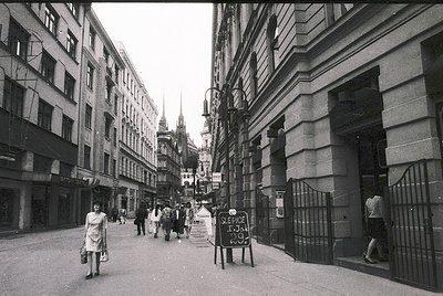Narrow urban street flanked by mid-rise Soviet-era buildings, likely Eastern Europe. Pedestrians in 1970s-era clothing; handw...