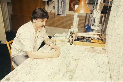 Mid-century workspace featuring a focused individual engrossed in detailed work on a large, flat surface. Surrounding materia...