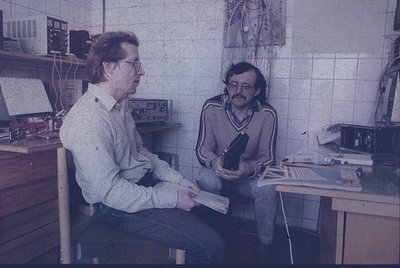 Two men in a retro office setting, likely mid-1970s–1980s. One holds a camera with a large lens, the other examines printed p...