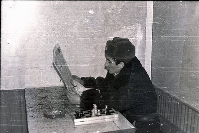 Vintage black-and-white photo of an elderly man in a peaked cap and heavy coat playing chess in an indoor setting, likely a p...