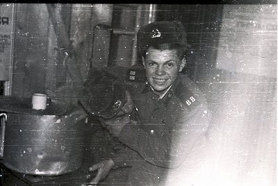 Young man in mid-20th century military uniform, likely Soviet-era, holding a large pot. Indoor setting with visible industria...