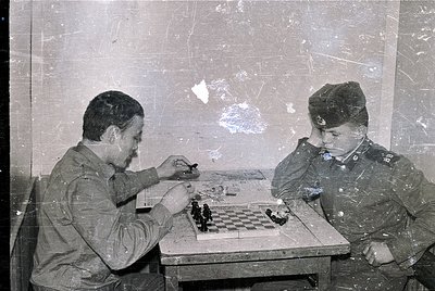 Two uniformed men play chess in a dimly lit, weathered room, likely mid-20th century. Uniforms suggest military or state serv...