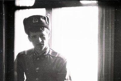 Young man in early 20th-century military uniform, likely 1910s–1930s, standing indoors near a window. Uniform includes peaked...