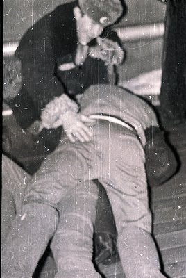 Black-and-white photo capturing a tense confrontation between two men in a dimly lit indoor setting, likely a bar or nightclu...