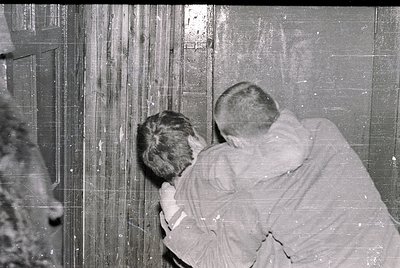 Two individuals in mid-20th century clothing examine a weathered, vertically striped wall—likely industrial or institutional....