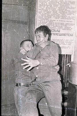 Intimate moment of a man hugging a young boy in an indoor setting, likely a public building. The Cyrillic text on the wall re...