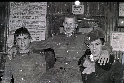 Three young men in Soviet-era military uniforms pose indoors, likely in a guard post or barracks. The man on the right wears ...