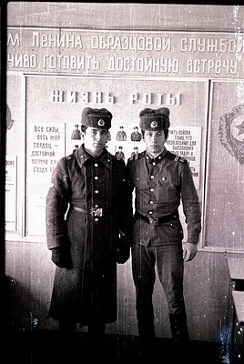 Vintage black-and-white photo of two young men in Soviet-era military uniforms, posed formally against a propaganda poster. T...