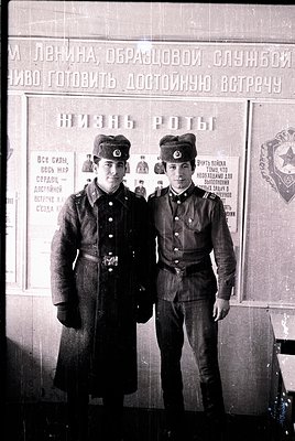 Two young men in Soviet-era military uniforms pose indoors, 1940s–1950s. Uniforms feature red star insignias, peaked caps, an...