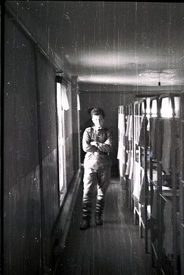 Mid-20th century industrial corridor with worker in overalls, standing near open doorway. Exposed pipes, bare lightbulbs, and...
