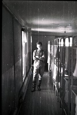 Mid-20th century man in uniform stands in a dimly lit, narrow hallway with rain streaks on windows. Industrial or institution...