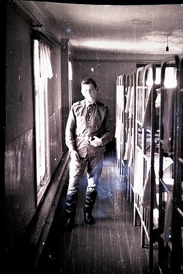 Young man in early 20th-century military uniform stands in a dimly lit corridor, holding a book or notebook. Wooden bunks lin...
